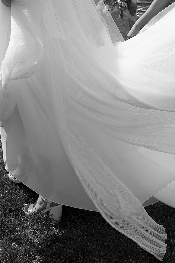 Wedding dress detail with pleated wedding dress overskirt flowing beside bridal heels and veil on a sunlit grass lawn outdoors