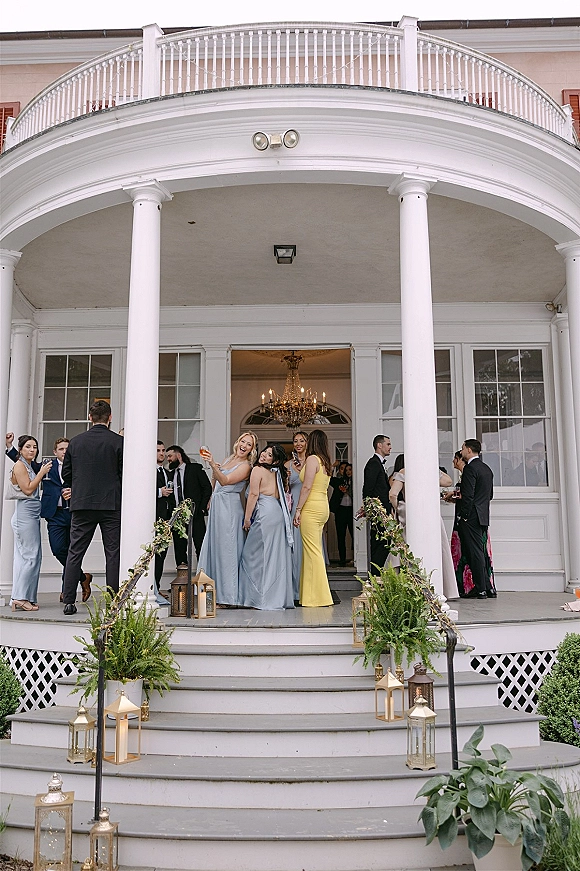 Wedding guests mingling at cocktail hour in formalwear, holding drinks on white columned porch steps with lanterns and greenery garland