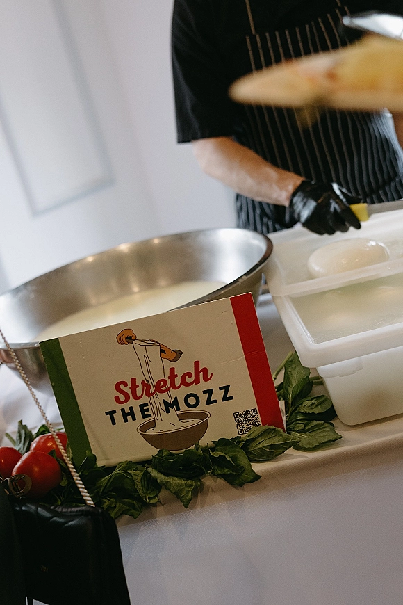 Mozzarella station with fresh mozzarella station curds and a ball in a metal bowl, gloved chef in striped apron beside basil and tomatoes signage with QR code indoors