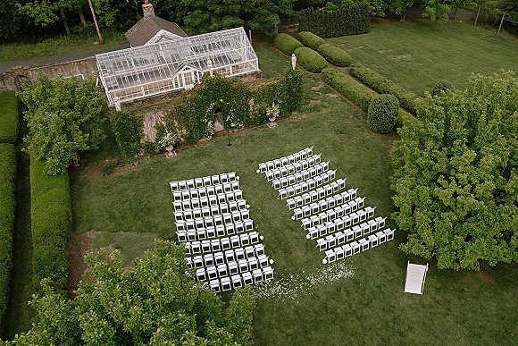 Outdoor ceremony setup with white chair rows and petal aisle leading to a simple arch on a lawn beside a glass greenhouse