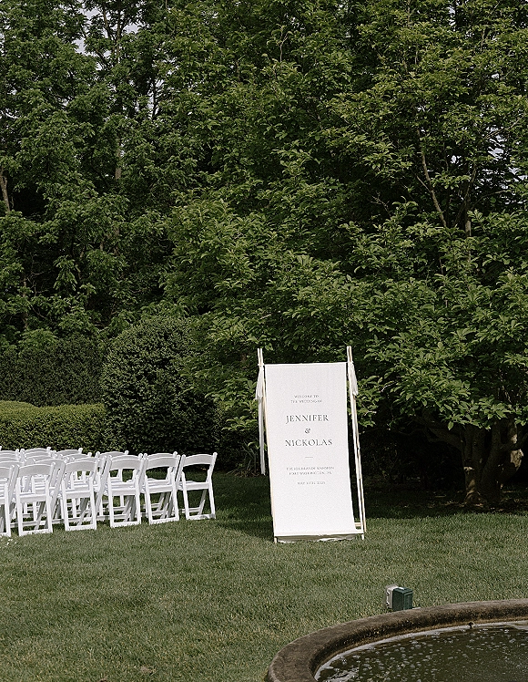 Wedding welcome sign on a wooden stand with ribbon ties, fabric panel, and white folding chairs on a green garden lawn by trees