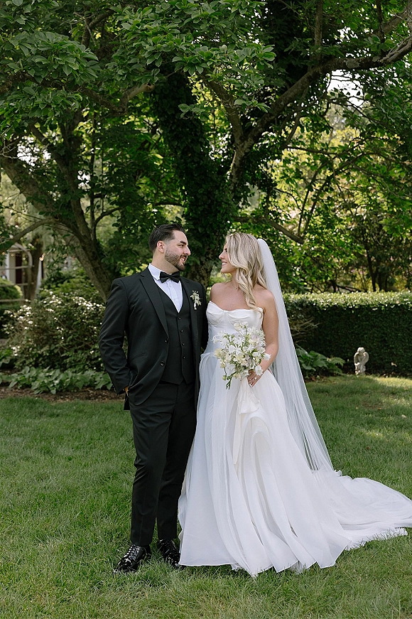 Couple portrait of bride in strapless dress with long veil and white bouquet beside groom in black tux on a garden lawn amid greenery