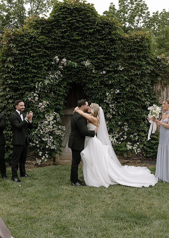 Wedding kiss portrait of bride and groom kissing, her long veil flowing as bridesmaids hold bouquets before a greenery wall and wooden door