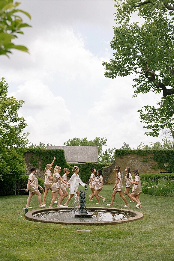 Bridal party photo of bride with bridesmaids in matching satin pajamas and slippers, holding champagne by a garden fountain on manicured lawn