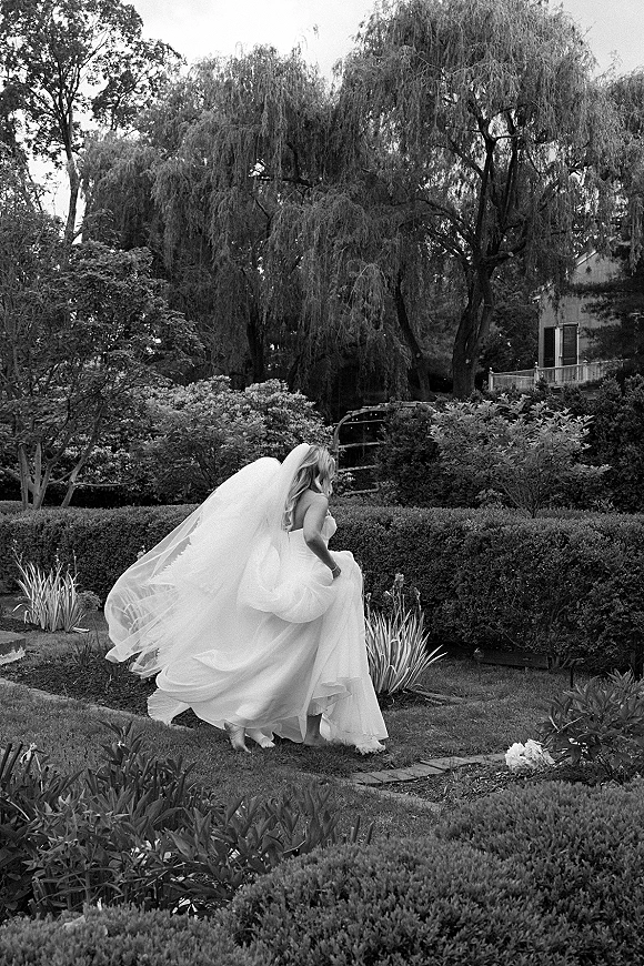 Bridal portrait of a bride running with veil flowing behind, lifting her full skirt on a stone garden path near hedges and house