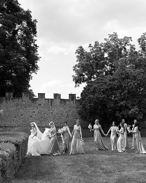 Bridesmaids portrait of the bride with bridesmaids holding hands, carrying bouquets on a garden lawn beside a stone wall under cloudy sky