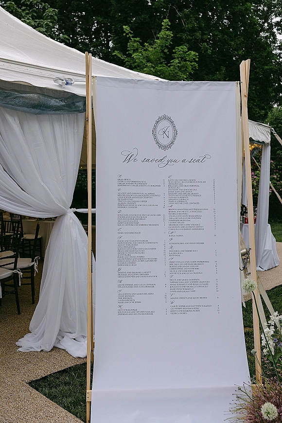 Wedding seating chart sign on fabric in a wood frame with monogram crest and calligraphy, accented by florals under an outdoor tent
