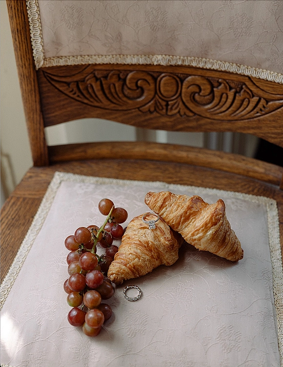 Wedding rings and engagement ring detail stacked on a croissant with grapes, styled on a neutral upholstered vintage chair seat