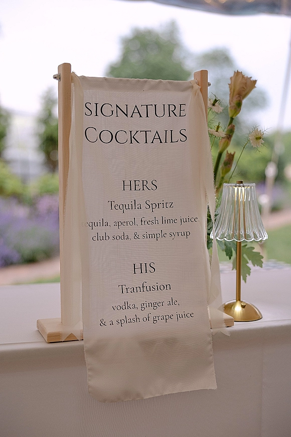 Wedding signature cocktail sign on a wooden stand beside a brass lamp and florals on a linen table in an outdoor garden tent setting