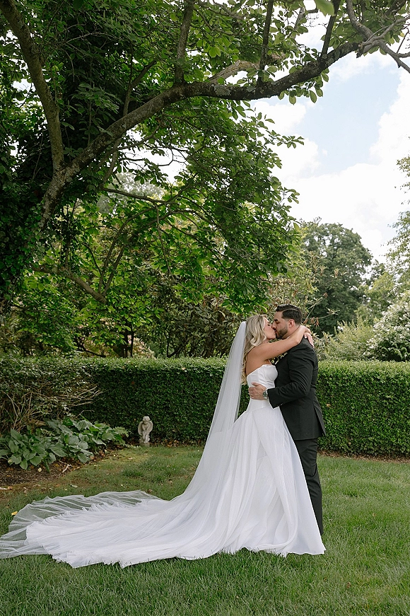 Wedding kiss portrait of bride and groom kissing, her long veil and tulle train flowing on a garden lawn beneath leafy trees
