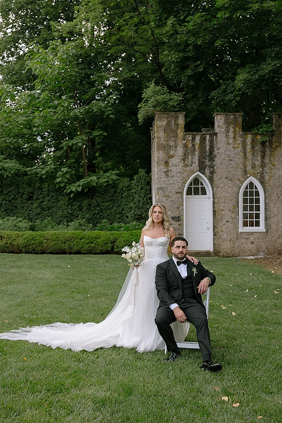 Couple portrait of bride standing with hand on seated groom’s shoulder, bouquet and long train before a stone building with arched windows