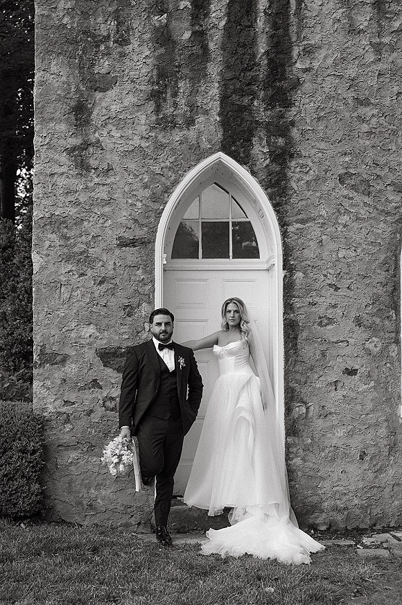 Couple portrait in a black and white wedding portrait style, bride in strapless dress and long veil beside groom in tux by arched doorway