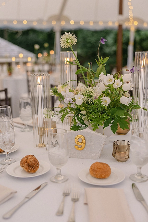 Reception tablescape with wedding table centerpiece, pillar candles in glass hurricanes, gold table number, and place settings under string lights in an outdoor tent