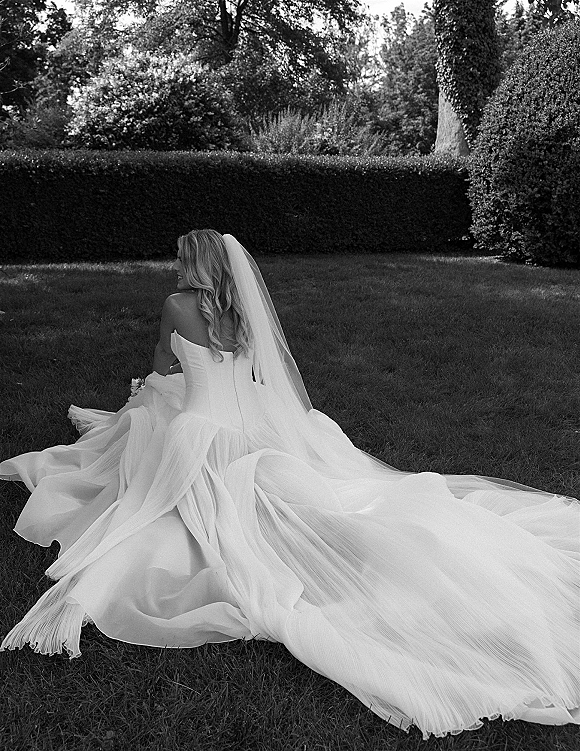 Bridal portrait in black and white of a bride sitting on grass, looking over her shoulder with a long train and veil in a garden