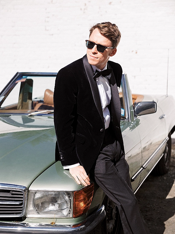 Groom portrait in a black tuxedo and bow tie wearing sunglasses, leaning on a vintage convertible car against a white wall