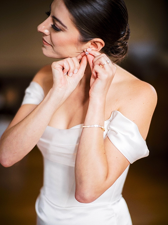 Bride getting ready, putting on earrings in an off-the-shoulder gown, pearl bracelet and wedding ring visible against a neutral wall