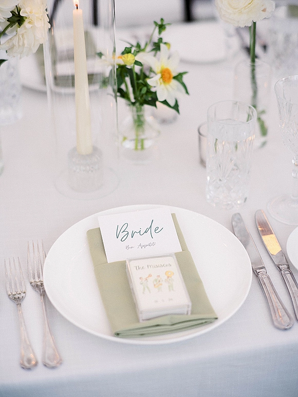 Reception place setting with wedding tablescape details: sage green napkin on white plate, menu and place card, candle and daisy bud vase on tablecloth