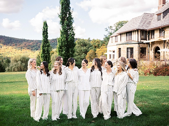 Bridesmaid group photo in matching pajama sets, laughing arm in arm on a lawn with trees, hills, and a stone manor house behind