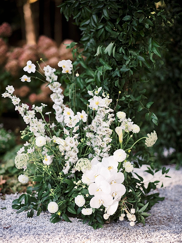 Wedding floral arrangement with white orchids and greenery, styled as ceremony aisle flowers along a gravel path beside garden shrubs