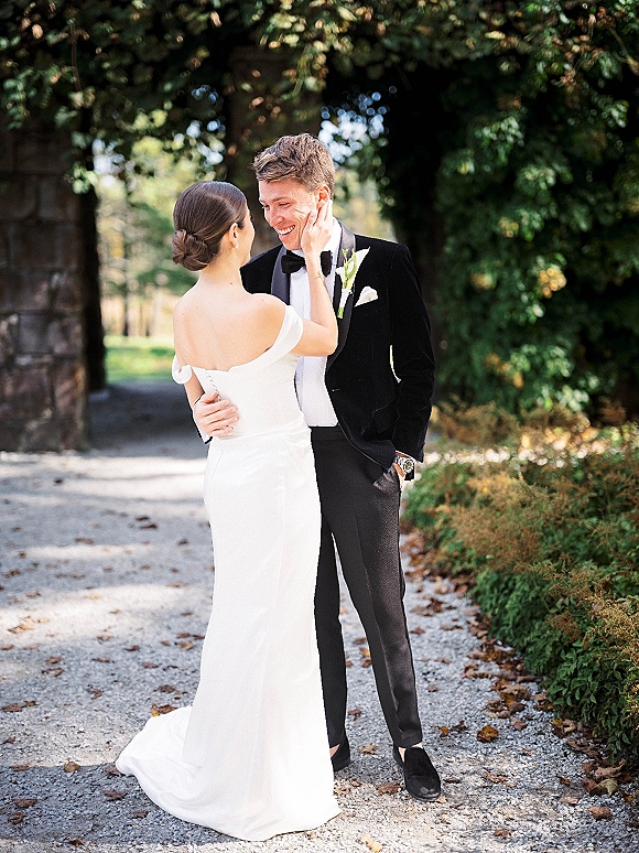 Couple portrait of bride touching groom’s face, both laughing, with off-the-shoulder gown and tuxedo on a tree-lined gravel path