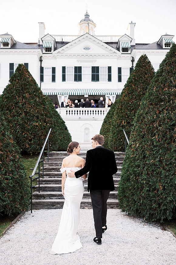 Couple portrait of bride and groom walking away in formal attire up stone steps at a white manor with balcony guests under striped awnings