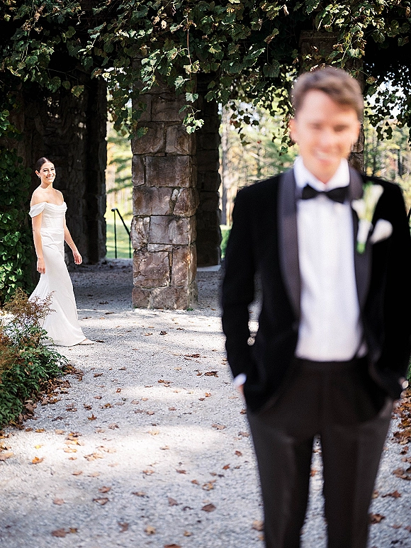Wedding first look as bride approaches groom in a tuxedo under an ivy-covered stone archway, smiling along a gravel garden path