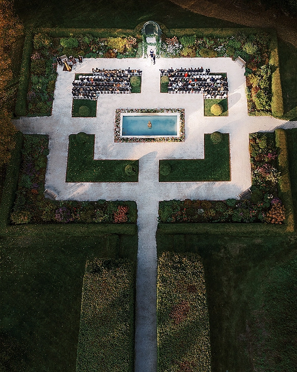 Outdoor ceremony setup in a formal garden wedding ceremony with white chair rows flanking a fountain, flower borders, arch, and string quartet