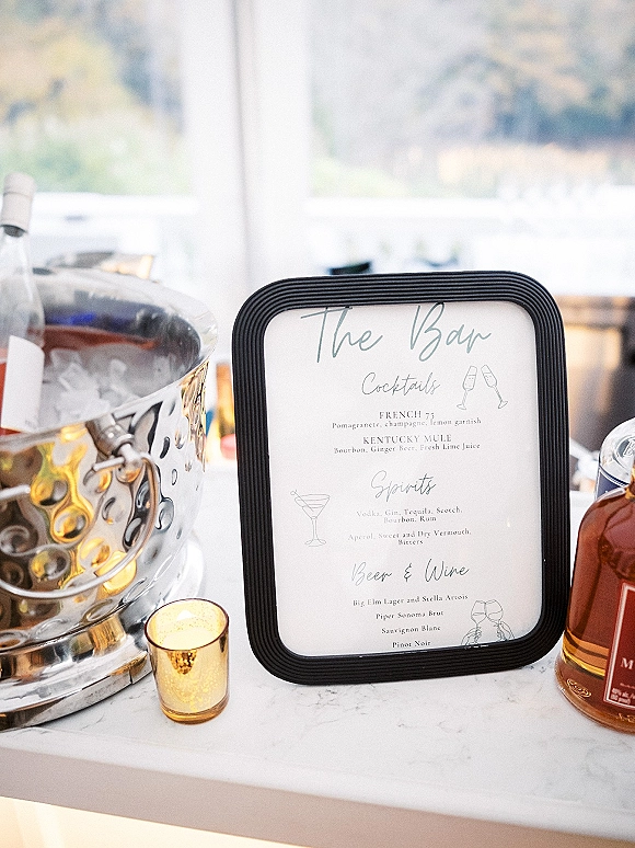 Wedding bar menu in a black frame with a wedding signature drink sign, champagne in an ice bucket and candlelight on a counter by a window
