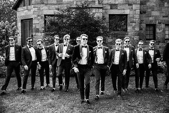 Groomsmen portrait with groomsmen walking photo style, wearing tuxedos, bow ties and sunglasses outside a stone building with shrubs and lawn
