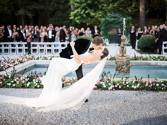 Wedding kiss portrait of bride in strapless gown as groom dips her, veil train flowing by reflecting pool fountain and garden chairs