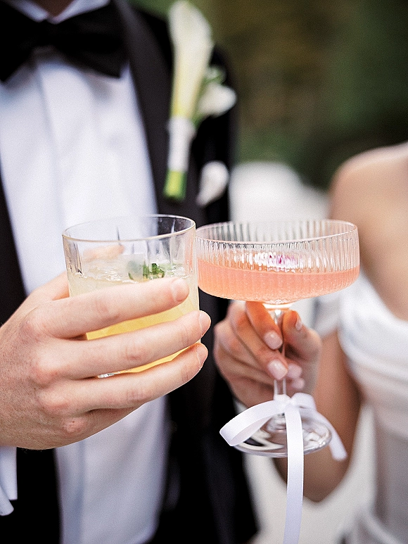 Wedding toast as bride and groom clink cocktail glasses, pink champagne in a coupe with ribbon bow, tuxedo and manicure against greenery