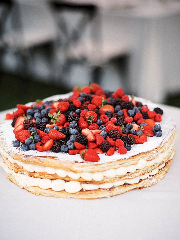 Wedding cake topped with fresh berries and powdered sugar, layered pastry with cream filling on a white tablecloth with blurred greenery background