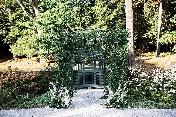 Wedding ceremony backdrop with a greenery ceremony arch on a lattice frame, white floral accents, set along a wooded garden path