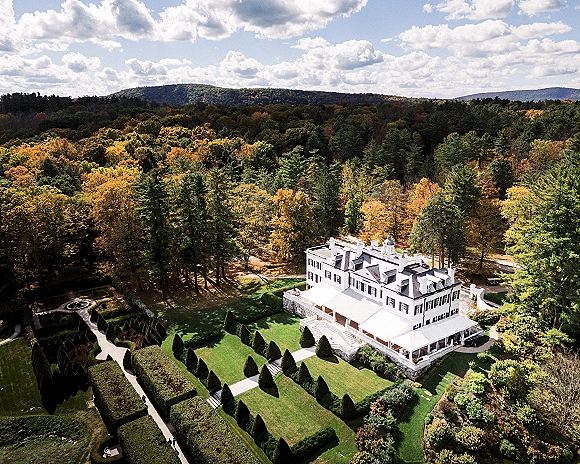 Wedding venue exterior showing a white manor house and formal gardens with topiary hedges, paths, and lawn, set against fall hills and forest