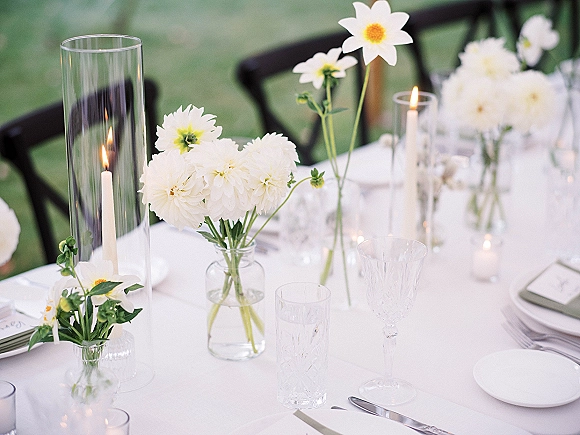 Reception tablescape with white wedding tablescape details—white florals in bud vases, taper candles in glass hurricanes, and classic place settings on an outdoor lawn