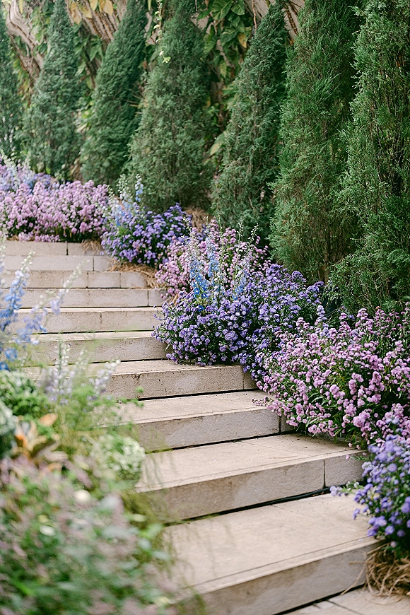 Ceremony aisle flowers with purple accents and greenery lining wooden steps, framed by evergreen shrubs and lush garden foliage outdoors