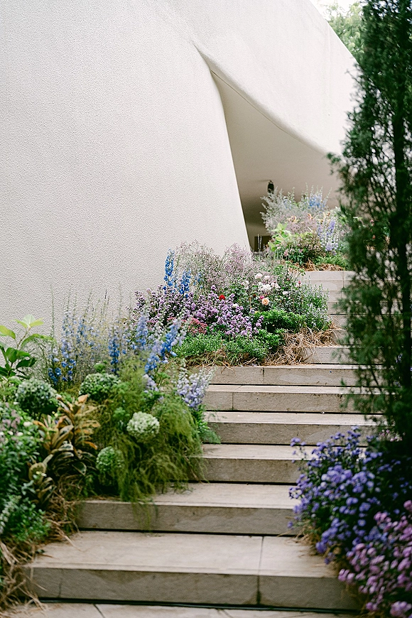 Ceremony aisle flowers and wildflower aisle decor line stone steps with purple and blue blooms against a white stucco curved wall