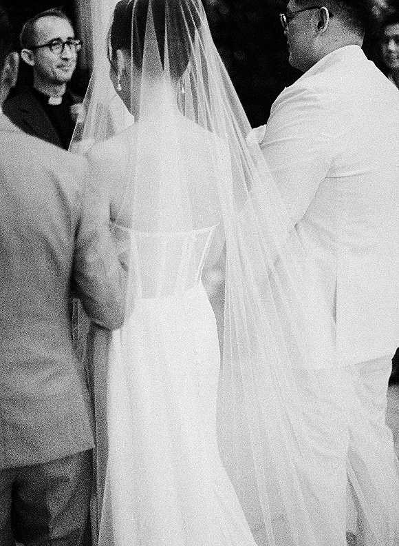 Ceremony moment as bride and groom at altar hold hands, veil flowing behind strapless dress while officiant speaks outdoors amid greenery