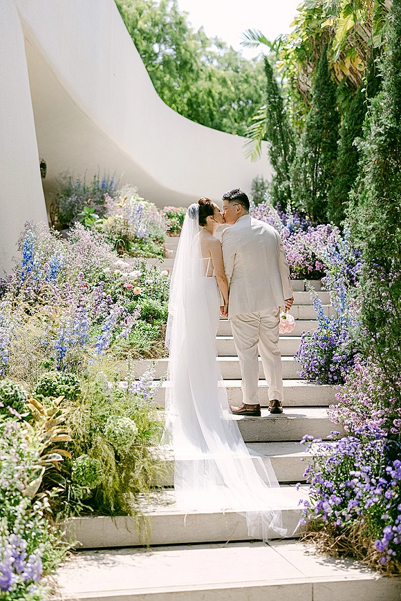 Wedding kiss portrait of bride and groom kissing from behind on garden steps, her veil flowing over a strapless dress beside wildflowers