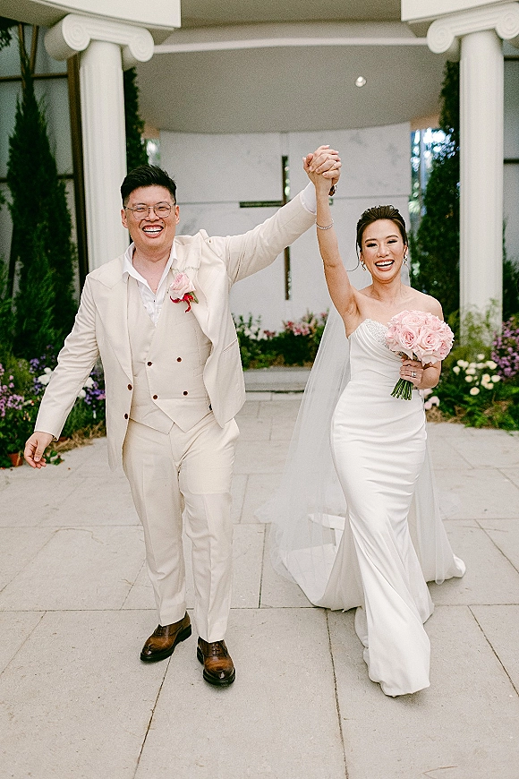 Couple portrait of newlyweds walking hand in hand, smiling with arms raised, bride in strapless gown and veil holding blush roses outside columns