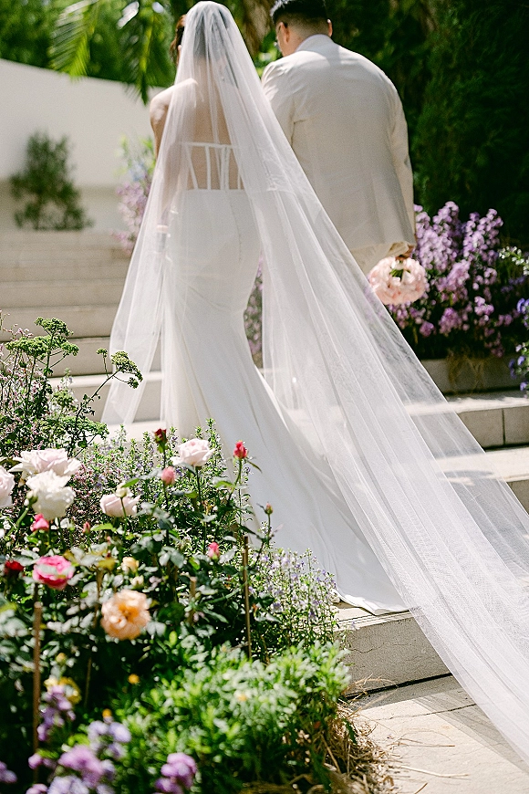 Wedding couple walking away, bride and groom from behind with a cathedral veil and bouquet on stone steps in a lush garden walkway
