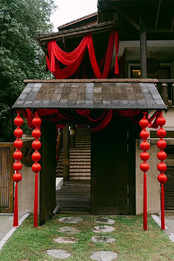 Wedding entrance decor with ceremony entrance draping in rich red fabric, hanging lanterns and tassels on a wooden gate by stone steps and lawn