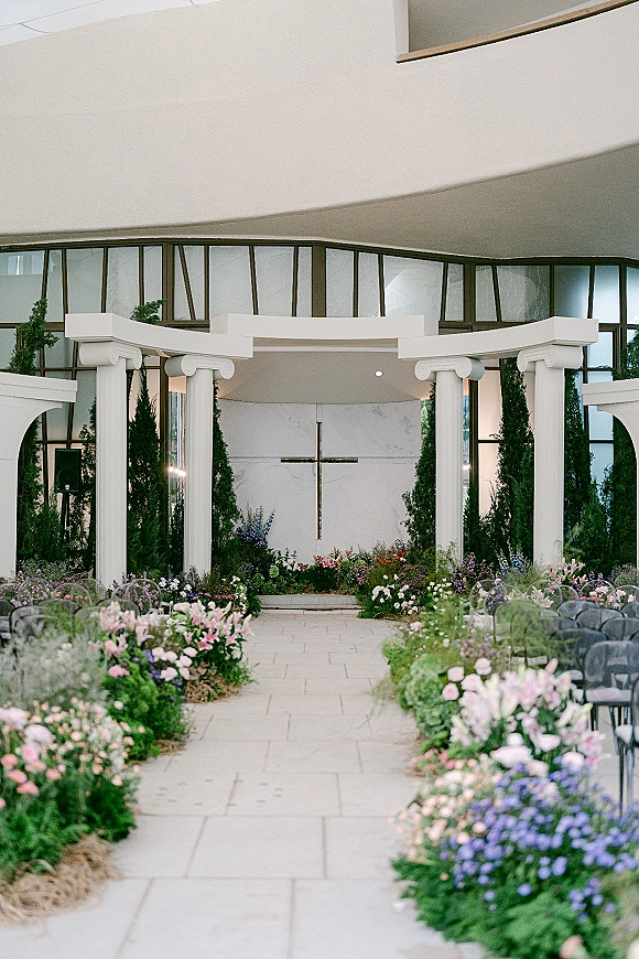 Ceremony aisle decor with floral lined wedding aisle, greenery and white columns leading to a cross altar in an arched indoor chapel