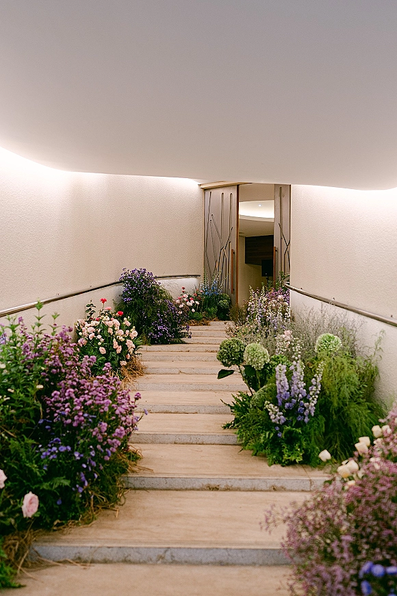 Ceremony aisle decor with wildflower arrangements and hydrangeas lining stone steps, set against curved white walls and wooden doors indoors
