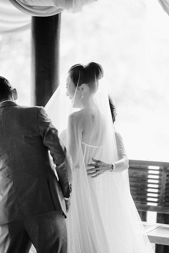 Ceremony moment as a bride walks down the aisle in a long veil and strapless dress toward a groom in a dark suit by bright window light