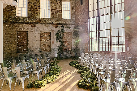 Ceremony setup with greenery aisle runners and metal chairs, set in a sunlit brick loft with large grid windows and lake view