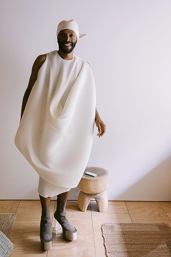 Groom portrait in a modern groom outfit, wearing a white pleated look with head wrap and platform boots against a neutral wall indoors