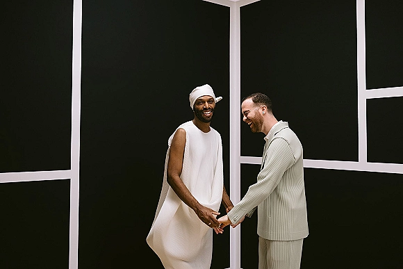 Couple portrait of a same sex wedding couple laughing and holding hands, one in a white headscarf, against black geometric wall panels