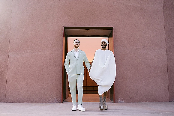 Couple portrait of a same sex wedding couple holding hands by an architectural doorway, one in a pastel green suit and loafers, the other in a white draped outfit with headscarf and platform boots