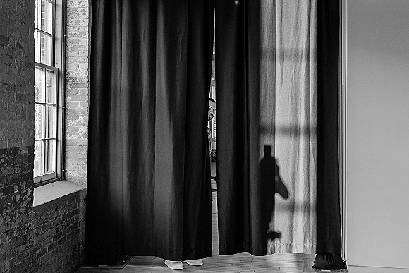 First look moment as groom peeks through black drapes at hidden bride, white bridal shoes visible by sunlit brick wall window
